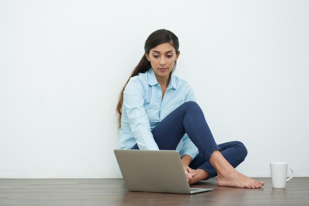 Serious Young Woman with Laptop and Tea on Floor