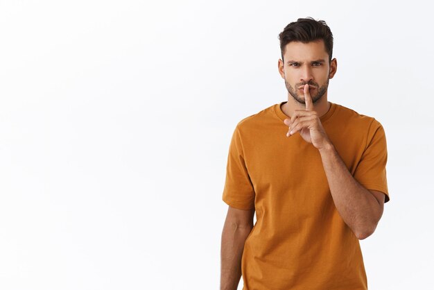 Seriouslooking concentrated and strict young man with beard shushing at camera with index finger pressed to folded lips frowning hushing demand stop talking loud say quiet please white background