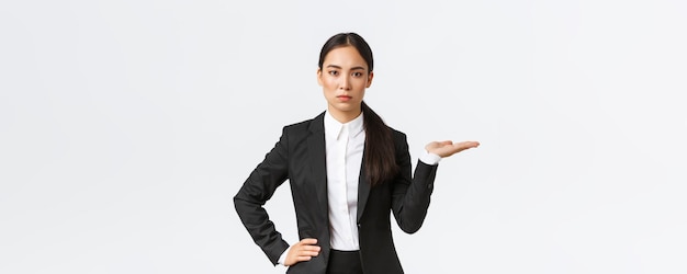 Seriouslooking determined young asian businesswoman introduce her project showing something on palm holding hand right on blank space as introduce product standing in black suit white background