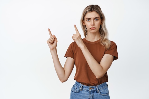 Free photo seriouslooking young woman pointing finger at upper left corner showing smth important making announcement standing against white background