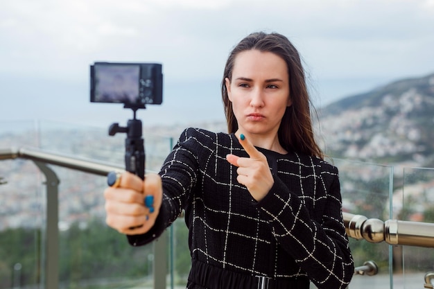 Seriously blogger girl is taking selfie by pointing camera focus with forefinger against the background of city view