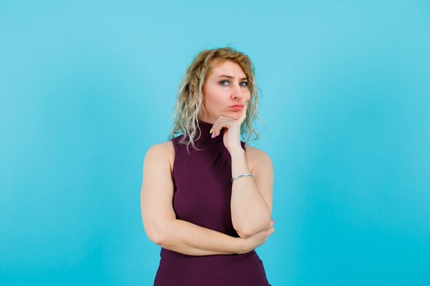 Seriously blonde woman is looking at camera by putting hand on chin on blue background