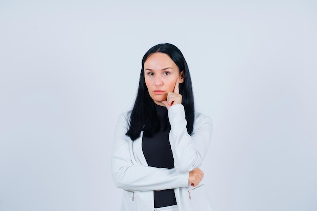 Seriously girl is looking at camera by holding forefinger on cheek on white background