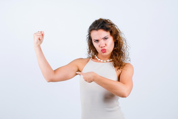 Seriously girl is raising up her muscle and showing it with forefinger on white background