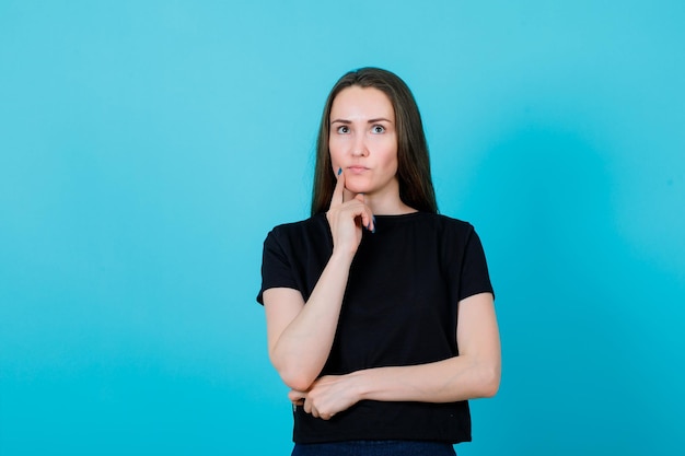 Seriously girl is thinking by holding forefinger on cheek on blue background