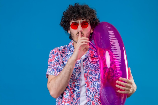 Seriously looking young handsome curly man wearing sunglasses holding swim ring gesturing silence on isolated blue space with copy space