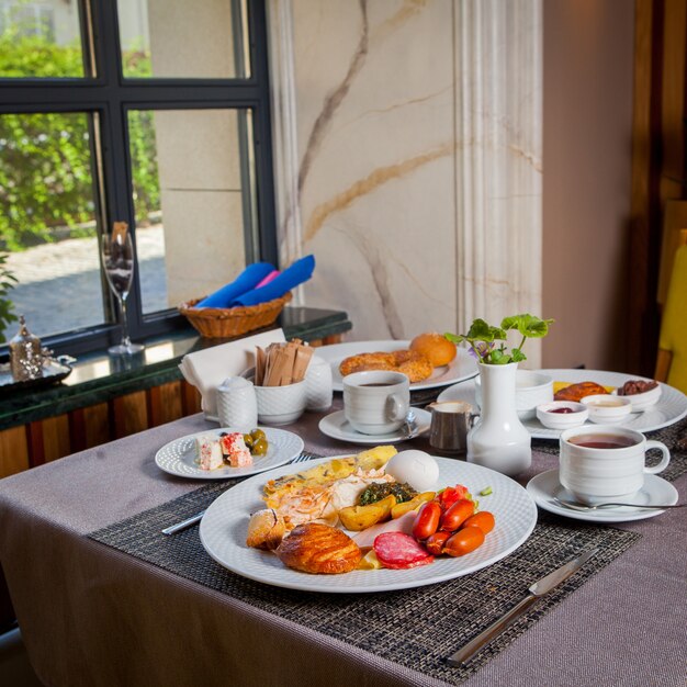 Served breakfast sausages, boiled egg, omelette, croissant in plates and cup of tea on table