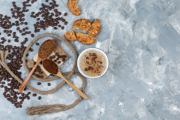 Set of cookies, coffee beans, grinded coffee, rope and coffee in a cup on grey plaster and wood piece background. top view.