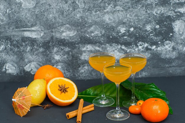 Set of juice in glasses, lemon, leaves, mandarin orange and oranges on dark and textured surface. side view.