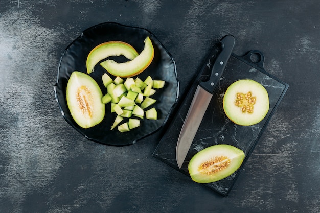 Set of melon and knife and sliced melon in a black bowl on a dark wooden background. flat lay.