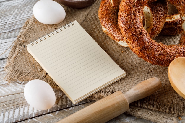 Set of notepad, eggs, rolling pin and turkish bagel on a sack cloth and wooden surface. high angle view.