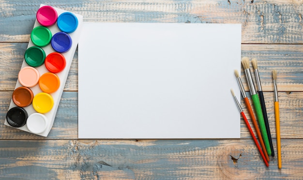 Set of water colors and paint brush with empty blank white paper over old wooden table