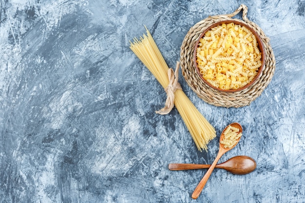 Free Photo set of wooden spoons and assorted pasta in a bowl on grey plaster and wicker placemat background. top view.