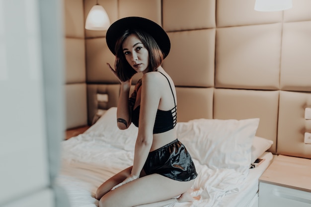 Sexy beautiful woman in fashion black underwear and wide hat sit on the bed in the morning