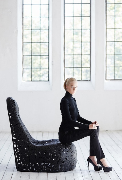 Free Photo sexy blond female in a black costume sits on a round black chair in  empty room.