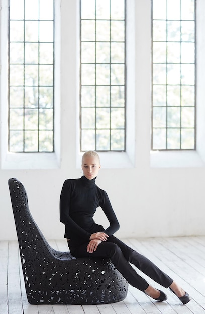 Free photo sexy blond female in a black costume sits on a round black chair in  empty room.