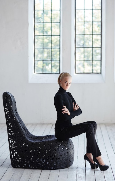 Free Photo sexy blond female in a black costume sits on a round black chair in  empty room.