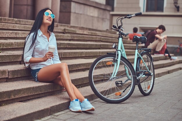 A sexy brunette female wearing blouse and   denim shorts in sunglasses, relaxing after riding on a bicycle, enjoying coffee on steps in a city.