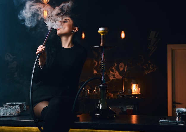 Free Photo sexy brunette girl in seductive black clothes smokes a hookah while sitting on counter in a nightclub.