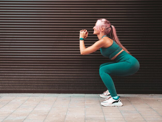 Sexy fitness woman in green sports clothing with pink hairShe doing squats Young beautiful model with perfect bodyFemale in the street near roller shutter wallCheerful and happy Outdoors