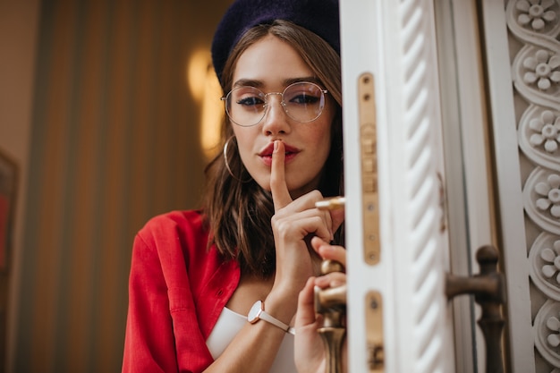 Free photo sexy lady with brunette hair, red lipstick in beret, shirt, stylish accessories posing and holding index finger to lips, looking at front and opening white doors