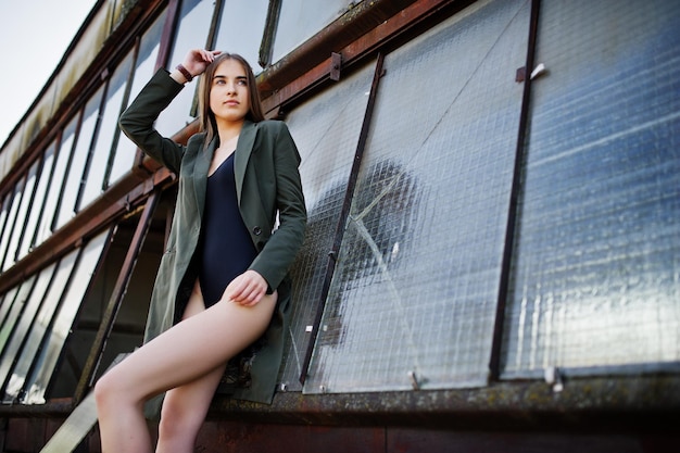 Sexy model girl wit long legs at black lingerie outfit body swimsuit combidress and jacket posed at the roof of abadoned industrial place with windows