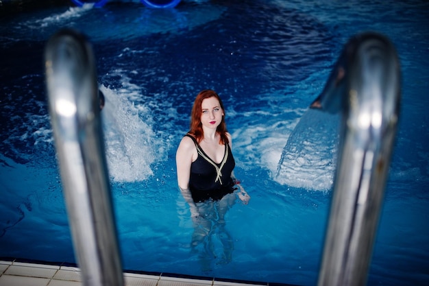 Sexy red haired girl in black swimsuit having rest in swimming pool of aqua park