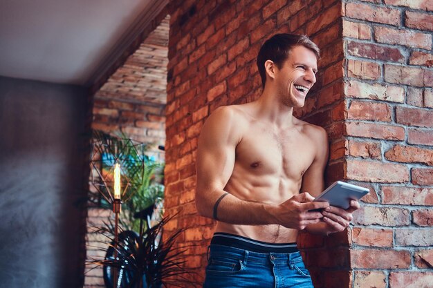 A sexy tattoed shirtless man with touchpad leans against the brick wall. Smiling and looking away.