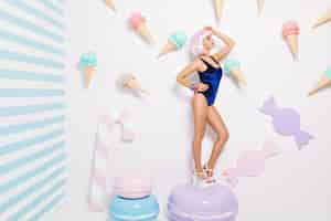 Free photo sexy young woman in blue bodysuit, on heels, with pink cut hairstyle standing on big macaron among sweets. joyful model, relax, sweet lifestyle, closed eyes.