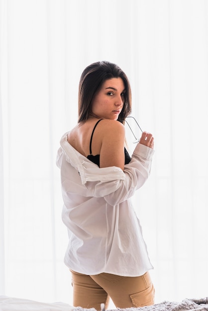 Sexy young woman holding eyeglasses in hand standing in front of curtain