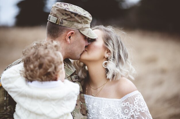 Shallow focus shot of an American soldier carrying his child while kissing his loving wife