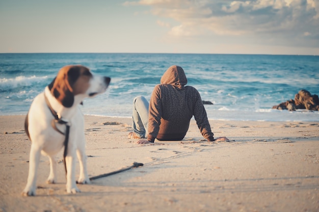 Shallow focus shot of a male in a black jacket sitting on a sandy beach near a beagle dog