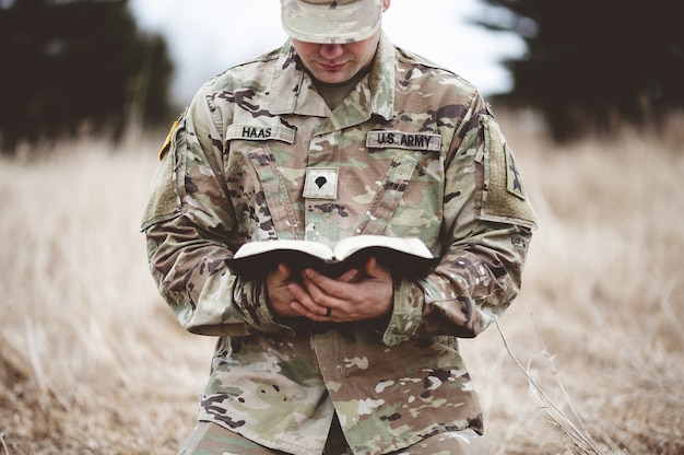 Shallow focus shot of a young soldier kneeling on a dry grass while reading the bible