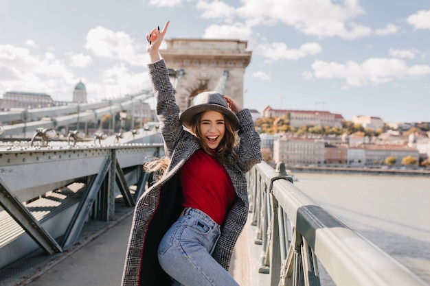 Shapely blissful girl in red sweater dancing on bridge on blur city background in autumn morning