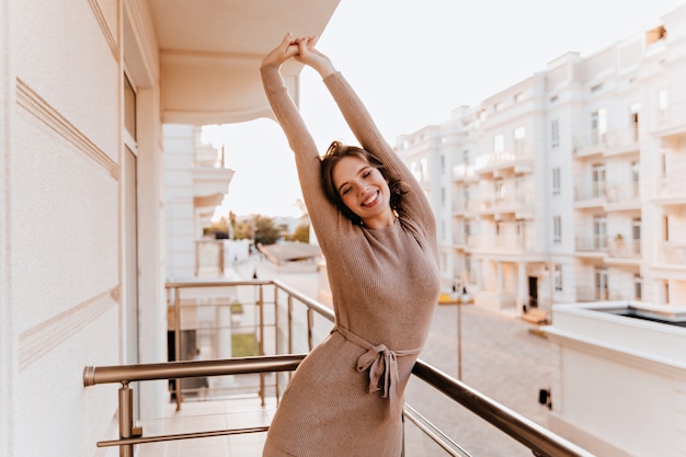 Free photo shapely caucasian woman in brown dress stretching at balcony. dreamy brunette girl enjoying morning at terrace.