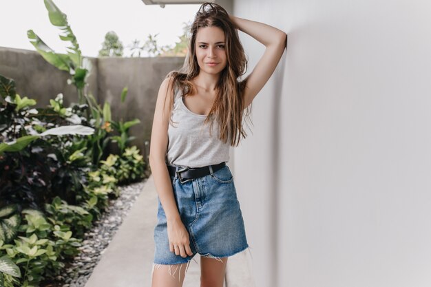 Shapely female model in summer attire standing in confident pose near white wall. Photo of serious brunette girl wears denim skirt looking