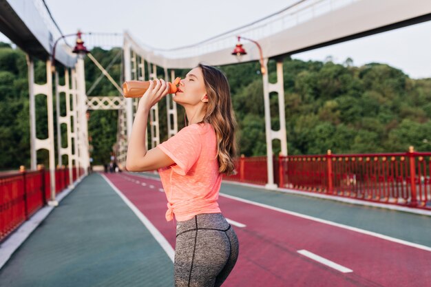 Shapely lady drinking water during marathon. Outdoor portrait of happy female runner standing at cinder track.