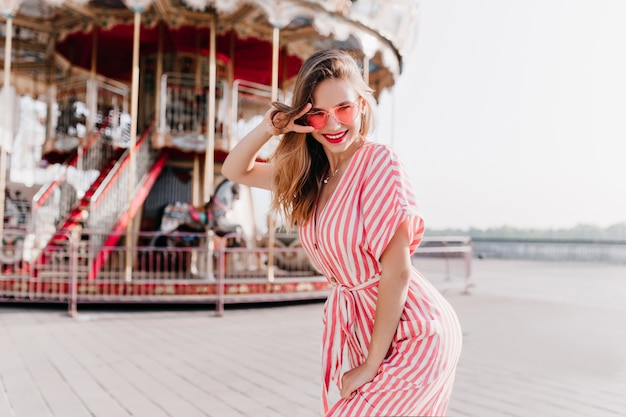 Shapely woman posing near carousel with inspired smile. Spectacular white girl in striped dress enjoying weekend in amusement park.