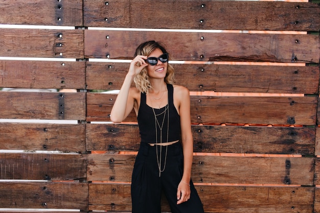 Free photo shapely young woman in black pants having fun during photoshoot. pleasant curly female model wears sunglasses and stylish tank-top.