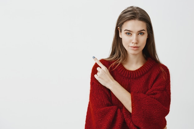 Sharing secret place with us. Confident good-looking female entrepreneur in red loose sweater, smiling with intrigued assured expression, pointing at upper left corner 