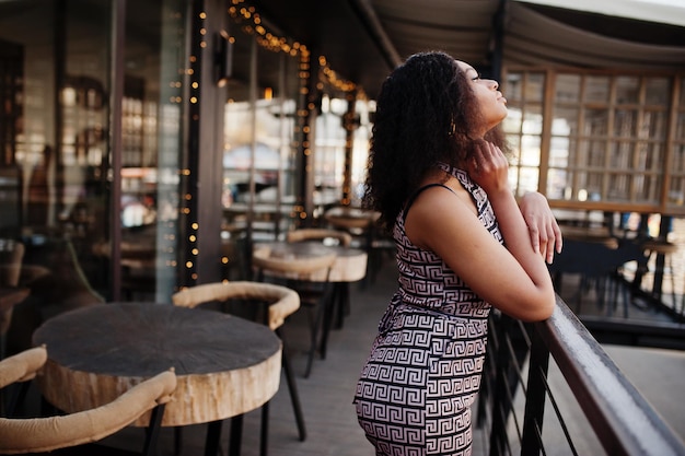 Free photo sharming elegant young african american woman with long curly hair wearing jumpsuit posing at cafe terrace outdoor against christmas decoration new year eve