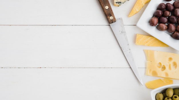 Free photo sharp knife with cheese and fresh olives on white wooden desk