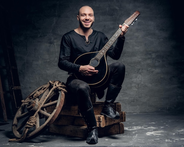 Shaved head traditional folk musician dressed in vintage Celtic clothes sits on a wooden box and plays mandolin.