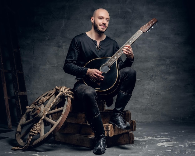 Free Photo shaved head traditional folk musician dressed in vintage celtic clothes sits on a wooden box and plays mandolin.