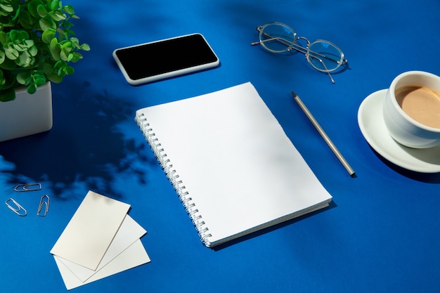 Sheets, coffee and work tools on a blue table indoors. Creative, cozy workplace at home office, inspirational mock up with plant shadows on surface. Concept of remote office, freelance, atmosphere.