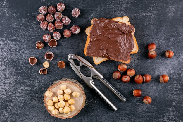 Free photo shelled hazelnuts with cocoa spread bread, nutcracker, piece of wood top view on a dark stone table