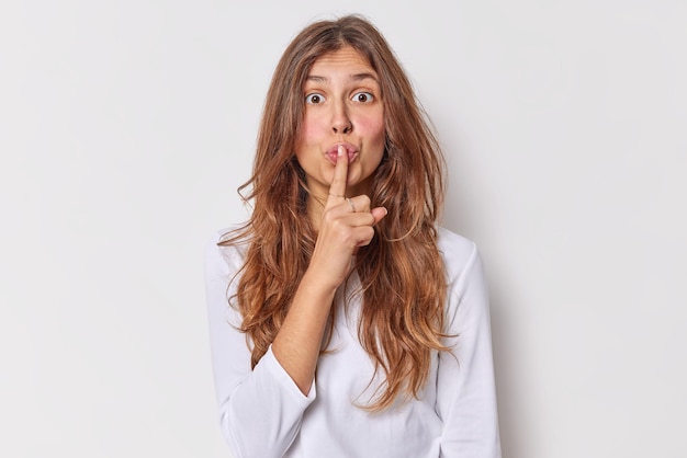 Free Photo shh its my secret. surprised long haired woman tells be silent keeps index finger pressed over lips makes shut up gesture dressed casually isolated over white background gossips and spreads rumors