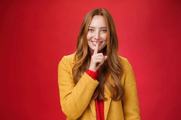 Shh keep it secret. Flirty and gentle attractive ginger girl with freckles teeling mysterious things showing shush gesture with index finger on mouth, smiling having surprise over red background.