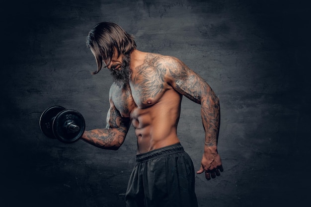 Free photo shirtless muscular, bearded male with a tattoo on his torso doing a biceps workout with dumbbell over dark grey background.