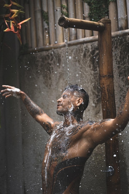 Free photo shirtless muscular latin american male with tattoos standing taking shower outloodrs after workout on the tropical leaves background. shower in the private villa.